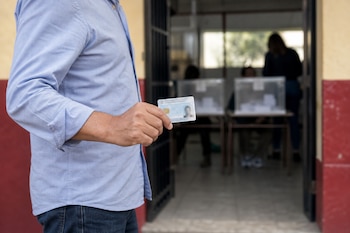 Un hombre sostiene una tarjeta de identidad frente a un recinto con mesas y urnas transparentes en el fondo.