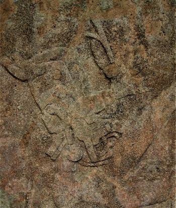 Grabado en piedra de color marrón con bajorrelieve que dibuja una figura humana con tocado y extremidades flexionadas