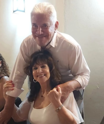 Hombre mayor de pelo blanco sonríe detrás de mujer mayor de pelo oscuro, ambos mirando a la cámara. Ella lleva una blusa blanca, él una camisa clara