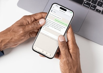 Manos de piel oscura sostienen un iPhone X plateado con pantalla mostrando un chatbot de IA sobre Microsoft; teclado de portátil difuminado al fondo