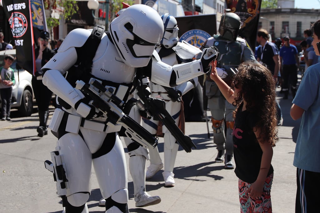 Una niña pequeña saluda a un Stormtrooper con armadura blanca en un desfile de Star Wars, creando un momento de alegría y conexión en la vía pública.