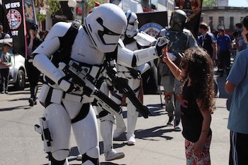 Un Stormtrooper de Star Wars, vestido con armadura blanca y negra, choca los cinco con una niña pequeña de cabello rizado en una calle soleada durante un evento
