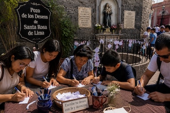 Personas sentadas a una mesa escriben en papeles con bolígrafos frente al cartel del Pozo de los Deseos de Santa Rosa de Lima y una estatua