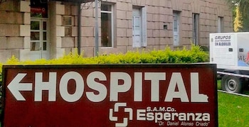 Un cartel marrón rojizo con letras blancas que dice "HOSPITAL Esperanza" y una flecha, frente a un edificio de tonos claros con ventanas y arbustos verdes