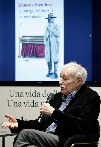 Eduardo Mendoza, un hombre mayor con pelo blanco y bigote, habla por un micrófono negro. Detrás, una pantalla muestra la portada de su libro