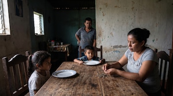 Una mujer sentada frente a una mesa de madera cuenta monedas. A su lado, una niña y un niño pequeños con platos vacíos. Un hombre de pie en el fondo.