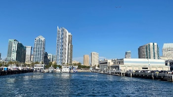 Imagen de un puerto con agua azul, barcos, muelles con coches y un horizonte urbano con rascacielos y edificios de gran altura bajo cielo despejado