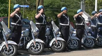 Una fila de policías motorizados uniformados, con cascos celestes, saluda firmemente mientras están de pie junto a sus motocicletas blancas de policía