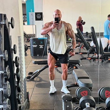 Un hombre musculoso con ropa deportiva toma una fotografía frente al espejo en una sala de pesas y bancos de ejercicio