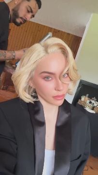 Una mujer con cabello rubio corto y ondulado, y vestida con una chaqueta negra, es peinada por un estilista en un interior, evocando la imagen de Marilyn Monroe. (belindapop/historias Instagram)