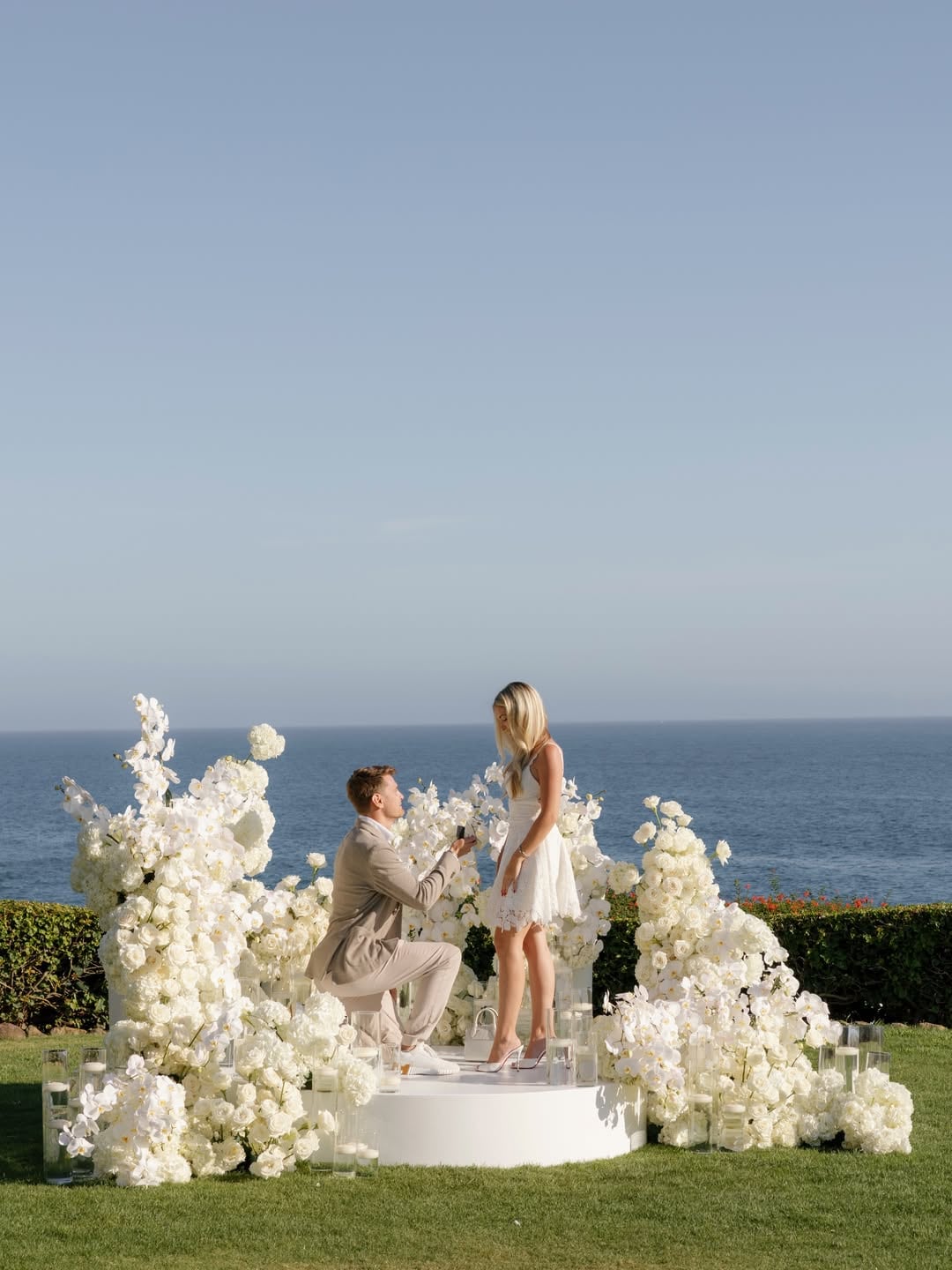 El piloto Logan Sargeant se arrodilla para pedir matrimonio a su pareja en una plataforma decorada con flores blancas frente al océano.
