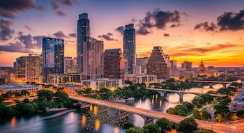 Vista aérea de la ciudad de Austin, Texas, al atardecer, con rascacielos altos, puentes iluminados sobre un río y un cielo naranja y morado.