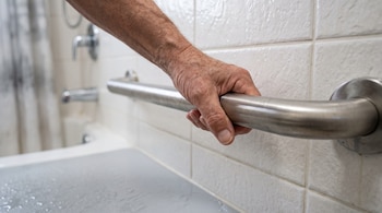 Una mano sujeta una barra metálica de apoyo instalada en la pared con azulejos blancos de un cuarto de baño.