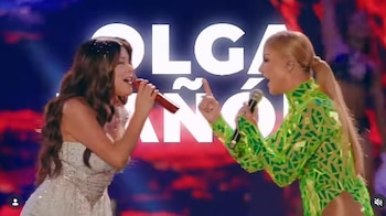 Dos cantantes femeninas en un escenario; una con vestido claro y micrófono rojo, la otra con atuendo verde y micrófono plateado, con texto "OLGA TAÑÓ" al fondo