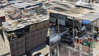 Vista aérea muestra dos bomberos con mangueras extinguiendo fuego en techo de un edificio con tejado de metal corrugado colapsado y barriles metálicos