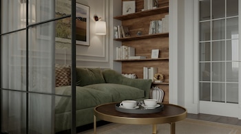 Vista de una sala de estar moderna con sofá verde, mesa de centro con dos tazas y estantería de madera llena de libros. Hay una partición de cristal a la izquierda.