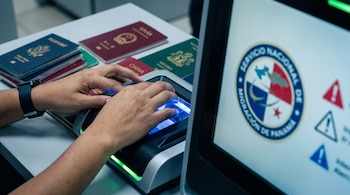 Manos sobre escáner de documentos con luz azul. Pasaportes de diferentes países en una mesa. Pantalla con escudo del Servicio Nacional de Migración de Panamá.