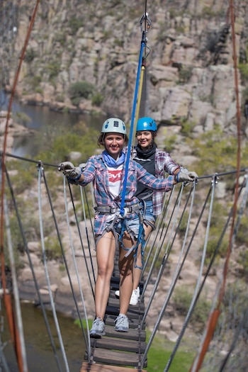 Dos mujeres sonríen con cascos, arneses y guantes mientras caminan por un puente colgante de madera y cables sobre un cañón con rocas y un río