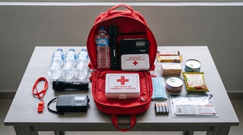 Mochila roja abierta sobre una mesa junto a botellas de agua, linternas, radios, botiquines de primeros auxilios, alimentos, pilas y documentos.