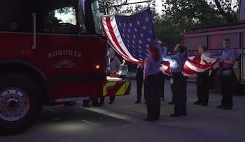Bomberos uniformados, varios hombres y mujeres, sostienen y despliegan una gran bandera de Estados Unidos junto a un camión de bomberos rojo con el texto "Augusta Fire Dept"