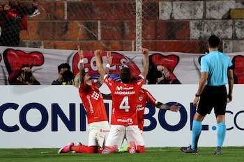 Tres futbolistas con camisetas rojas, dos arrodillados y uno de pie, celebran en el campo de juego. Un árbitro de espalda y aficionados se ven al fondo