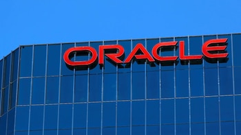 Primer plano del letrero rojo brillante de 'ORACLE' en la parte superior de un edificio de oficinas moderno con fachada de cristal azul reflectante bajo un cielo azul claro