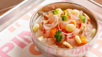 Tazón metálico con caldo, trozos gelatinosos translúcidos, semillas, aros de cebolla, dados anaranjados y hojas de cilantro