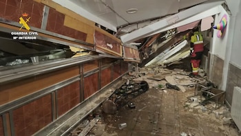 Interior de un local con el techo colapsado, escombros en el suelo y una persona con traje de emergencias al fondo