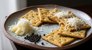 Varias galletas saladas cuadradas con queso rallado por encima y al costado, junto a un montón de granos de pimienta negra, servidas en un plato rústico.