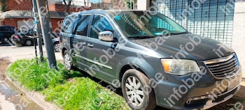 Minivan gris oscuro estacionada junto a una acera con pasto, un poste de luz, otros autos en segundo plano y edificios al fondo