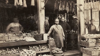 Fotografía en sepia de dos hombres en un mercado, uno con delantal, rodeados de puestos con tubérculos y carnes secas colgando