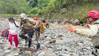 Un rescatista con casco blanco y ropa embarrada agarra la mano de una niña mientras caminan por un lecho de río rocoso. Otros rescatistas los siguen