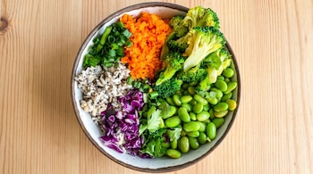 Vista cenital de un tazón de Buda con brócoli, zanahoria rallada, edamame, repollo morado, cebolla verde y granos sobre una superficie de madera clara.