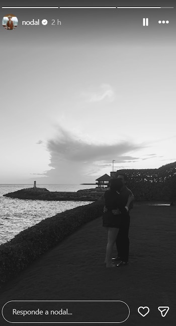 Fotografía en blanco y negro de dos personas abrazándose con el mar y un faro al fondo bajo un cielo nublado al atardecer