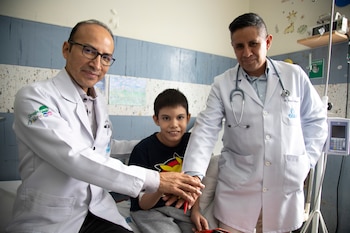 Dos médicos con batas blancas unen sus manos sobre la mano de un niño sentado en una cama de hospital