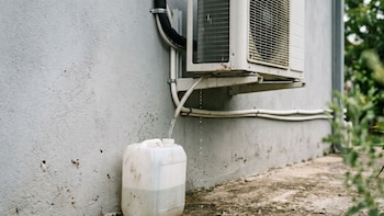 Unidad de aire acondicionado exterior montada en una pared gris, un tubo drena agua en un bidón blanco parcialmente lleno sobre el suelo