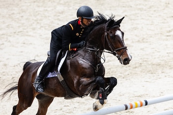 Jinete militar con uniforme negro y casco saltando con un caballo marrón sobre una valla en un campo de arena