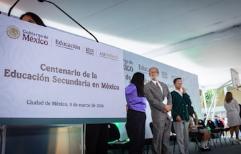 Há uma placa branca na frente anunciando o 'Centenário da Educação Secundária do México' com uma imagem do Governo do México. Várias pessoas estão participando de um evento em segundo plano