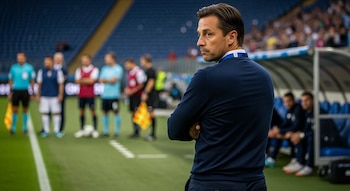 Un hombre con un suéter azul oscuro y los brazos cruzados está de espaldas al campo de fútbol, mirando de perfil, con jugadores y la tribuna desenfocados detrás.