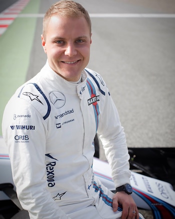 Primer plano de Valtteri Bottas sonriendo, con mono de carreras blanco y logos de patrocinadores, sentado junto a un coche de F1 en una pista