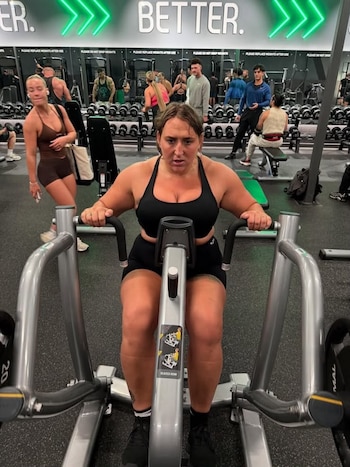 Una mujer sentada en una máquina de remo de gimnasio con ropa deportiva negra frente a pesas y espejos