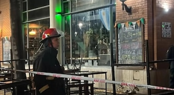 Un bombero con casco rojo de perfil junto a mesas exteriores de un local acordonado con una cinta de advertencia blanca y roja