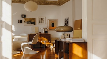 Interior de una sala de estar iluminada por el sol, con suelo de madera, una lámpara de diseño en un estante de madera y cuadros en la pared.
