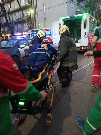 Bomberos y personal de emergencias trasladan a una persona en camilla hacia una ambulancia. Se observa una patrulla policial y edificios urbanos