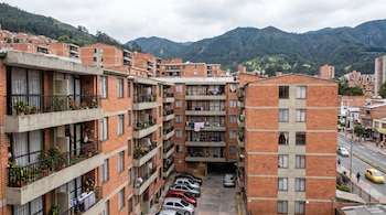Varios edificios de ladrillo con balcones y ropa tendida, un aparcamiento con coches, una calle con vehículos y peatones, y montañas de fondo.