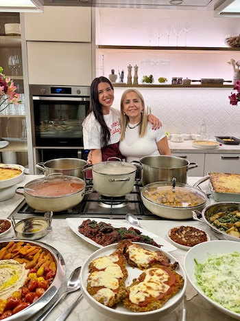 Dos mujeres posan en una cocina moderna detrás de una barra llena de fuentes de comida y ollas sobre los fogones