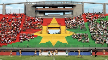 Ilustración de un estadio de fútbol con gradas repletas de aficionados en colores rojo, verde y una estrella amarilla. Varios jugadores en el campo de juego.