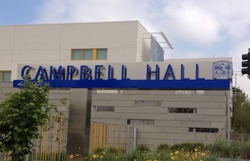 Letrero metálico con el nombre Campbell Hall en letras azules frente a un edificio moderno bajo un cielo nublado