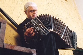 Primer plano de César Angeleri con cabello blanco, sosteniendo un bandoneón oscuro contra una pared ocre texturizada. Parte de una baranda negra es visible detrás