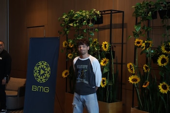 Um sorridente Louis Tomlinson, parado em frente a girassóis e folhagens, com uma bandeira azul escura do BMG à sua esquerda. Ele veste uma camiseta e jeans rasgados
